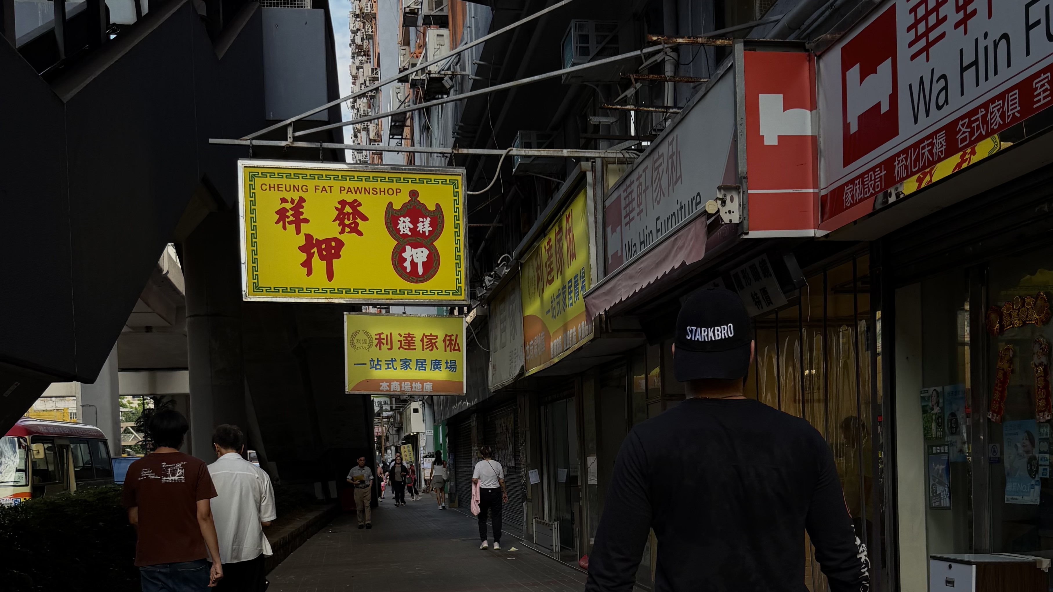 A person wearing a black STARKBRO cap walking through a Hong Kong street lined with signs